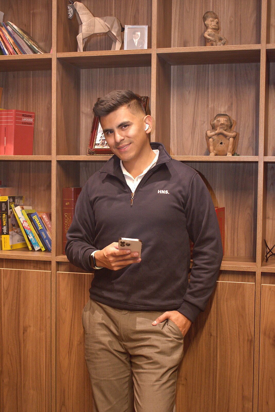 Johan Angel smiling in front of a bookshelf, holding his phone