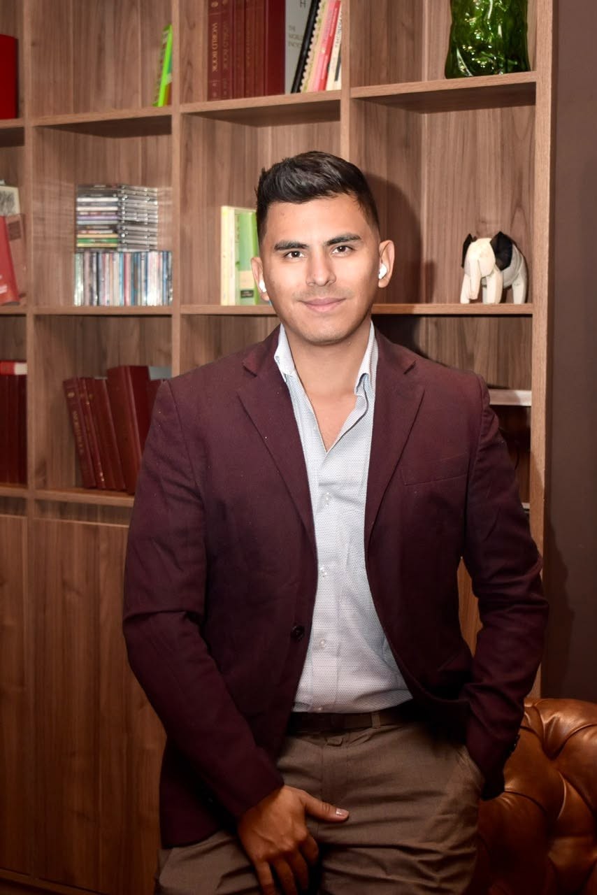 Johan Angel in a burgundy blazer, smiling in front of a wooden bookshelf