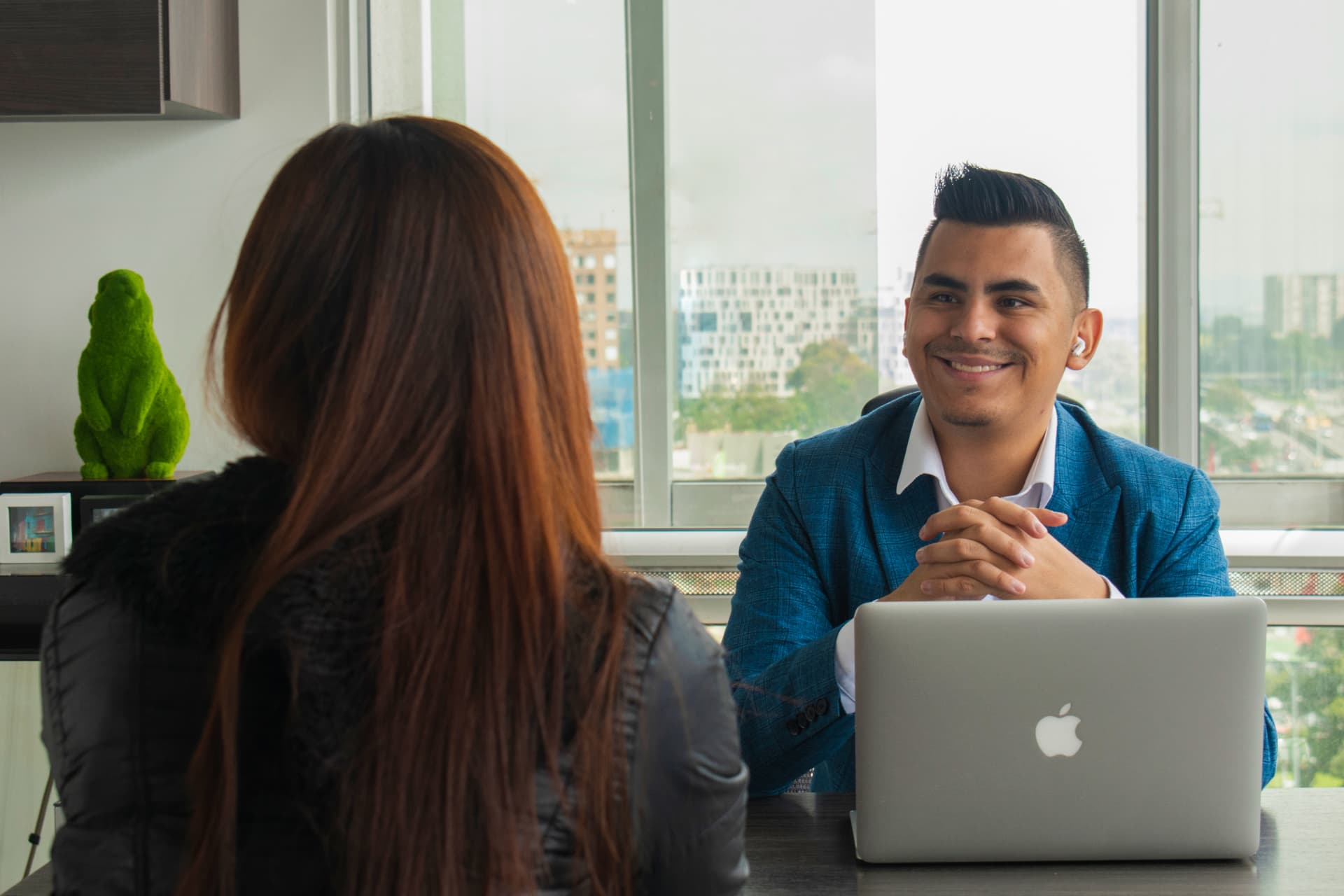 Johan Angel sonriendo en una reunión con una clienta en su escritorio, con la ciudad de fondo