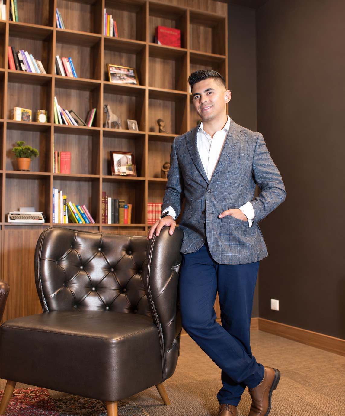 Johan Angel in a gray suit, standing next to a leather armchair in front of a bookshelf