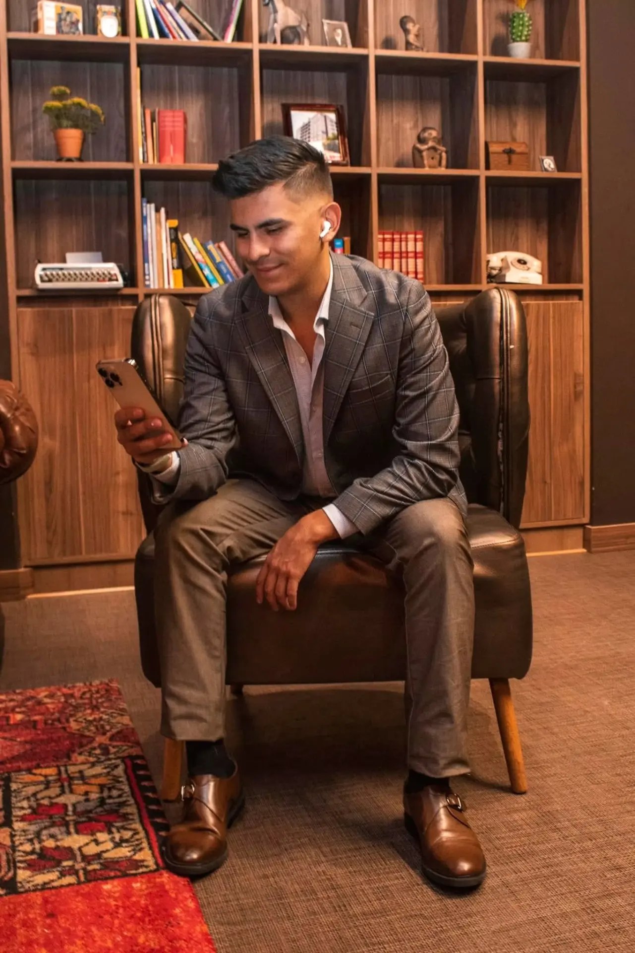 Johan Angel con blazer gris a cuadros, sonriendo mientras mira su teléfono en un sillón de cuero