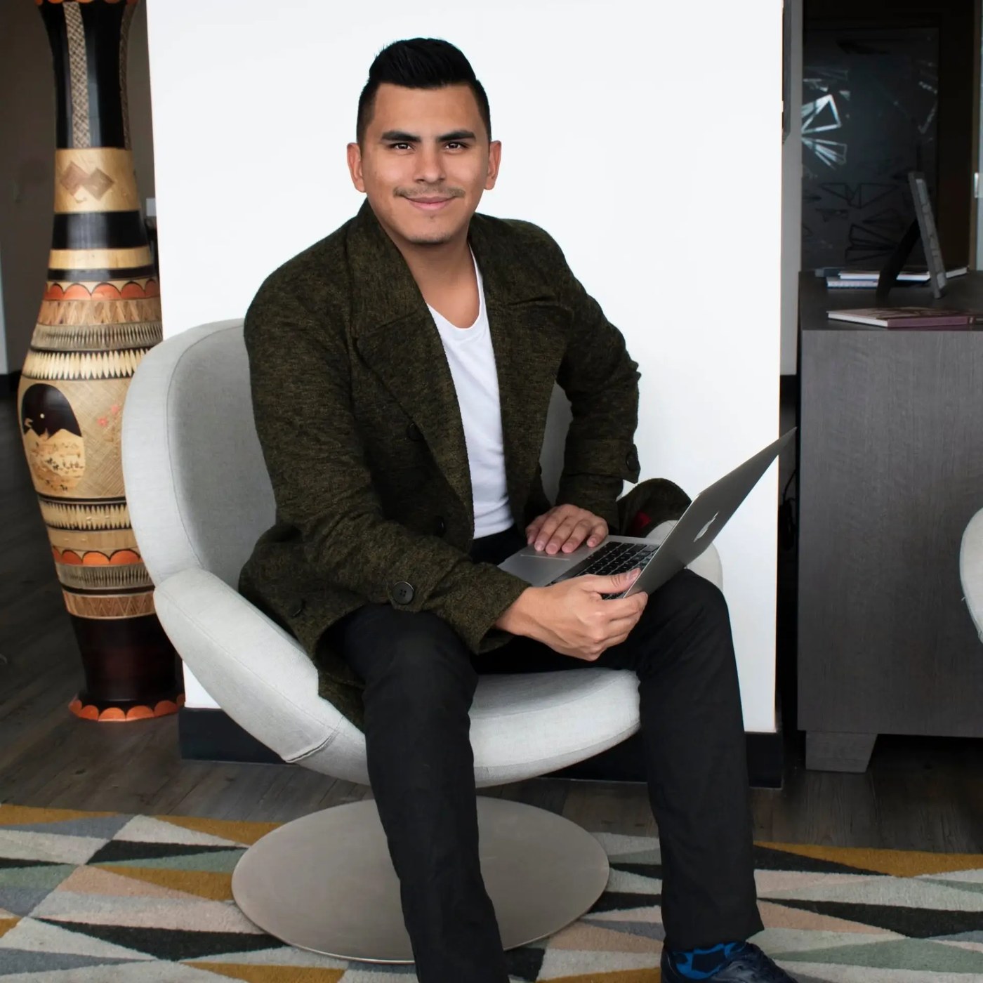 Johan Angel sonriendo en una oficina moderna, sentado en un sillón blanco con su portátil